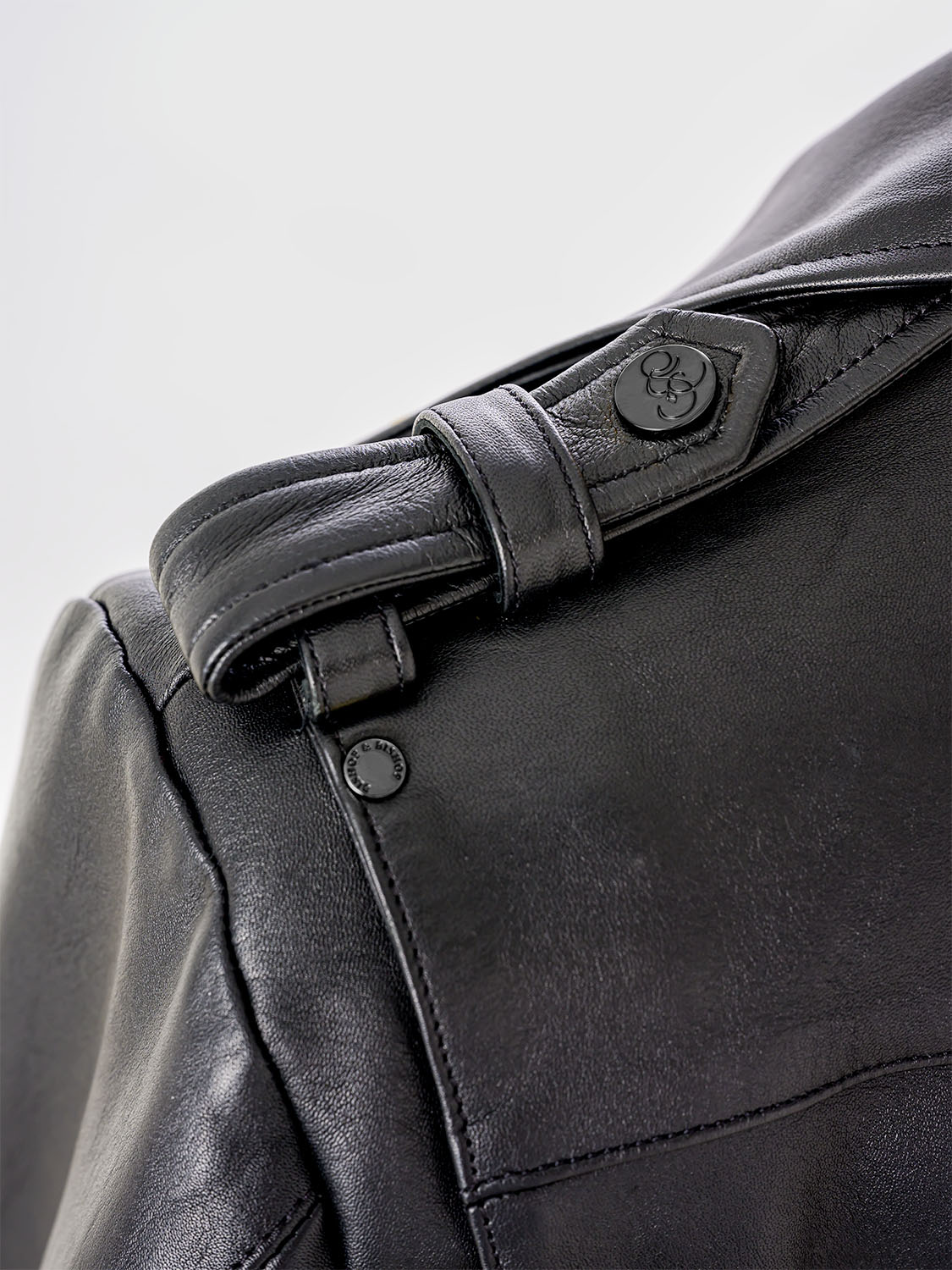 Close-up of the shoulder strap of a black leather jacket, showing a button engraved with the “Bishop & Bishop” logo.