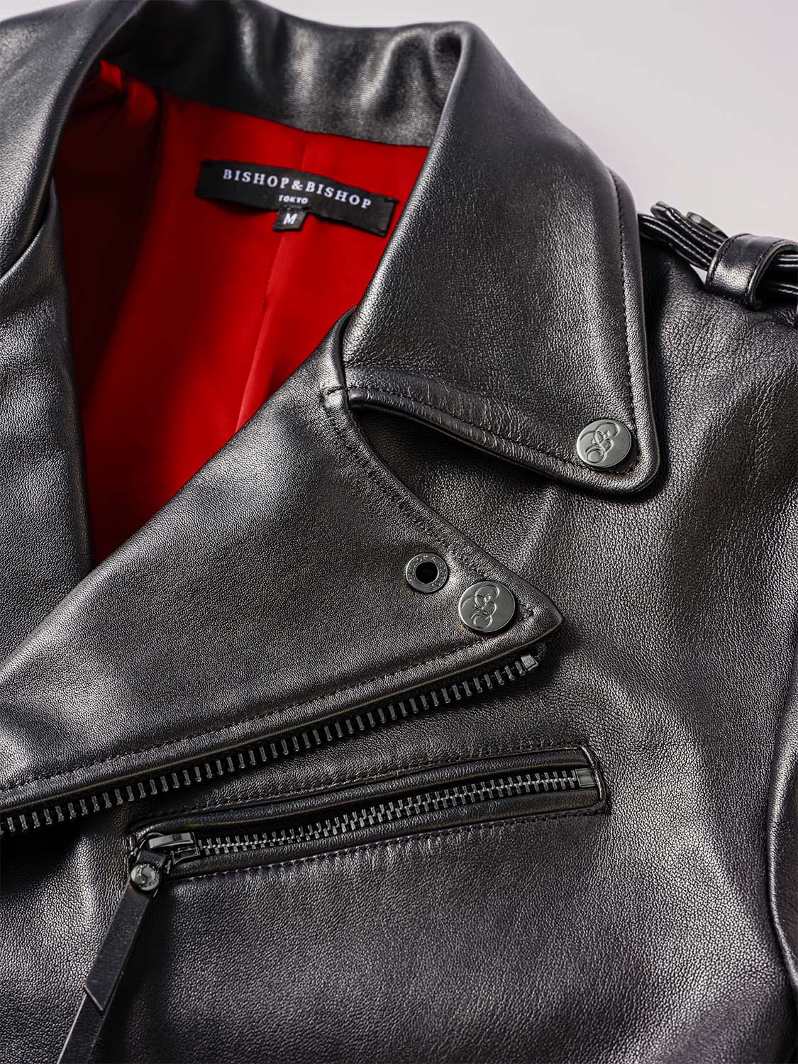 Close-up of the front collar of a black leather jacket, showing buttons engraved with the “Bishop & Bishop” logo and red interior lining.