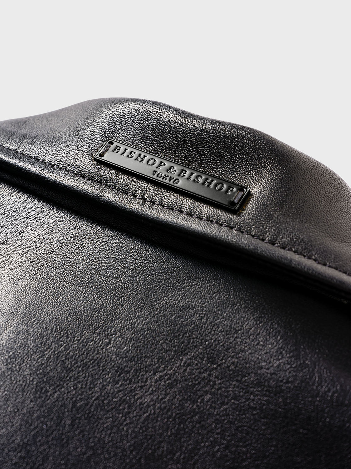 Close-up of the back of a black leather jacket collar, showing a black plate engraved with “Bishop & Bishop Tokyo.”