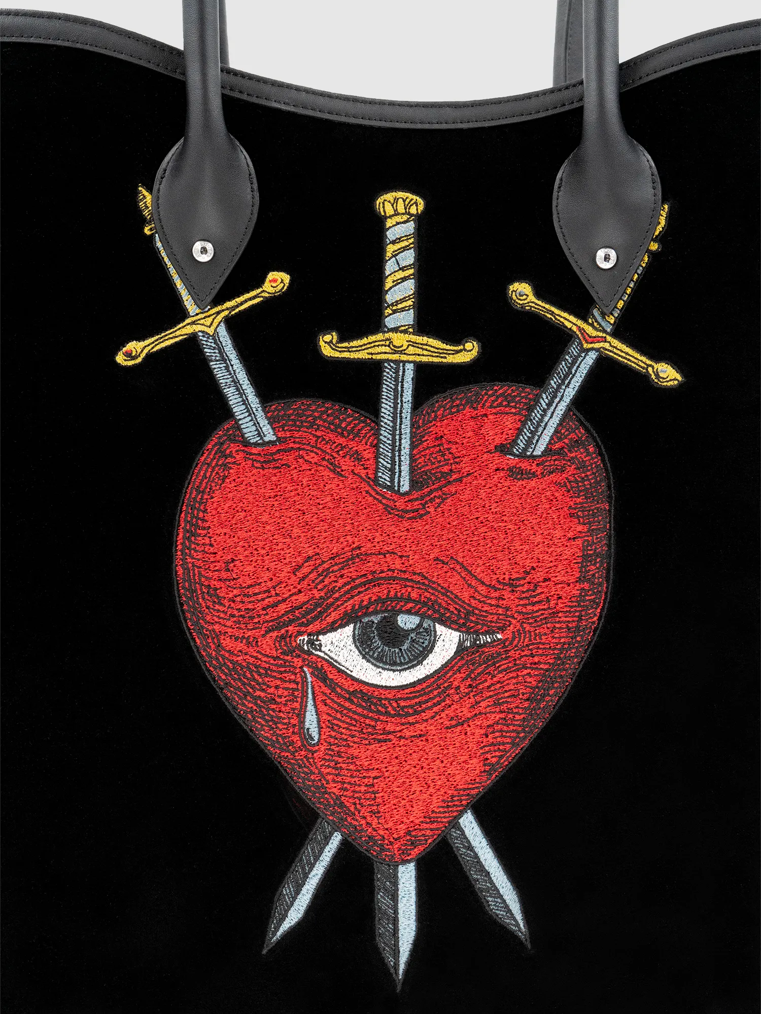 Close up of a black velvet and leather tote bag with a red heart, an eye, and crossed swords embroidered on the front, with a black handle.