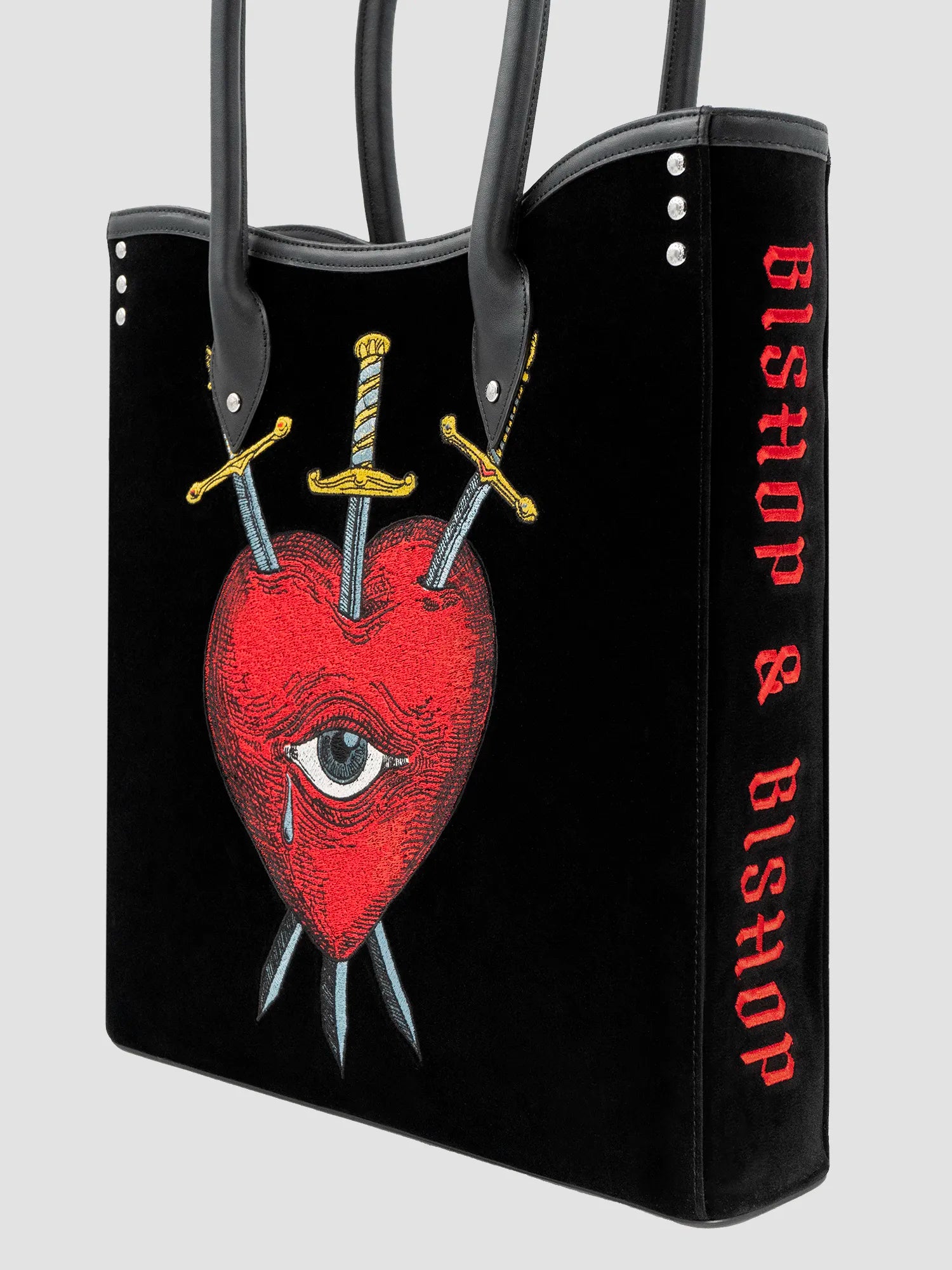 Side view of a black velvet and leather tote bag with a red heart, an eye, and crossed swords embroidered on the front, with a black handle.