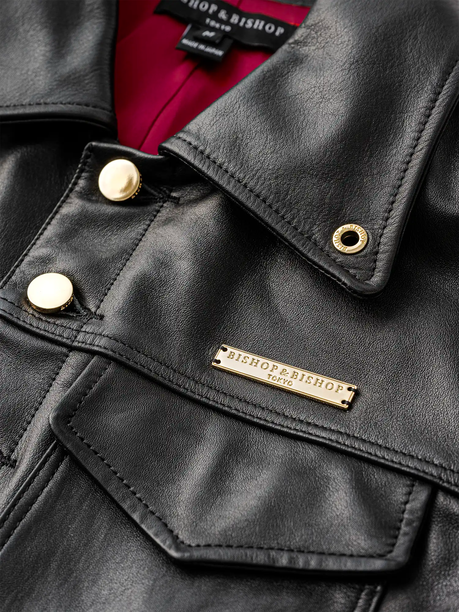 Close-up of the collar area of a black leather jacket with gold buttons and a gold plate engraved with “Bishop & Bishop Tokyo,” showing red interior lining.