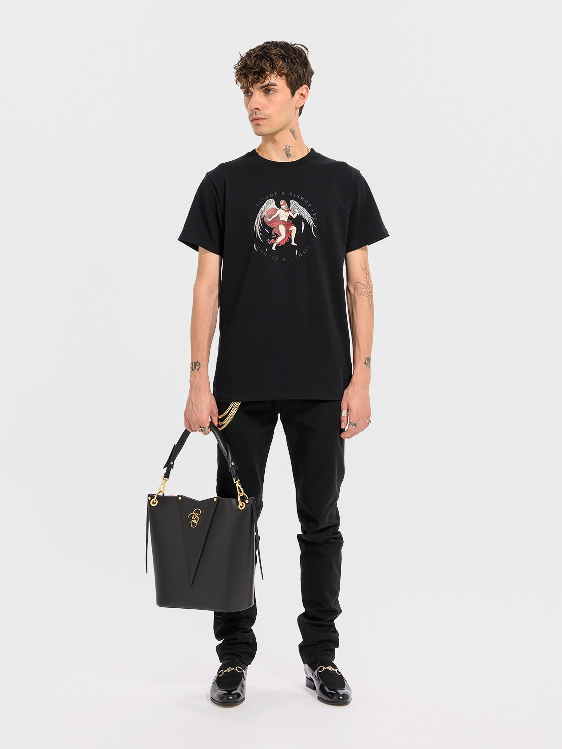Man wearing a black T-shirt featuring an angel with wings and a red blindfold graphic, holding a black handbag, on a white background.