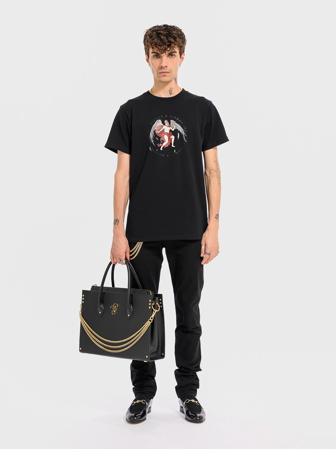 Person wearing a black t-shirt with a graphic design and holding a black leather handbag with gold-toned alloy parts, featuring a detachable shoulder strap and a brand emblem on the front.