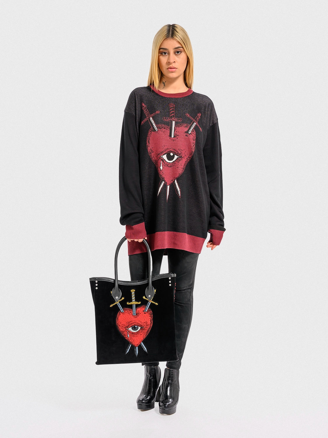 Person wearing a black sweater with a red heart, an eye, and crossed swords design, holding a matching handbag on a white background