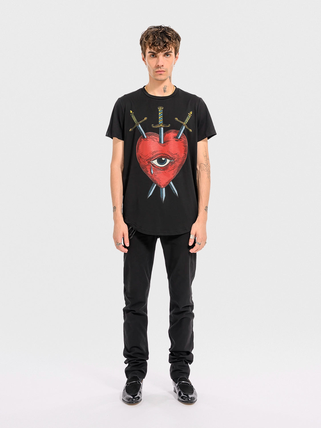 Person wearing a Black crew neck t-shirt with a graphic print of a red heart, an eye, and crossed swords.