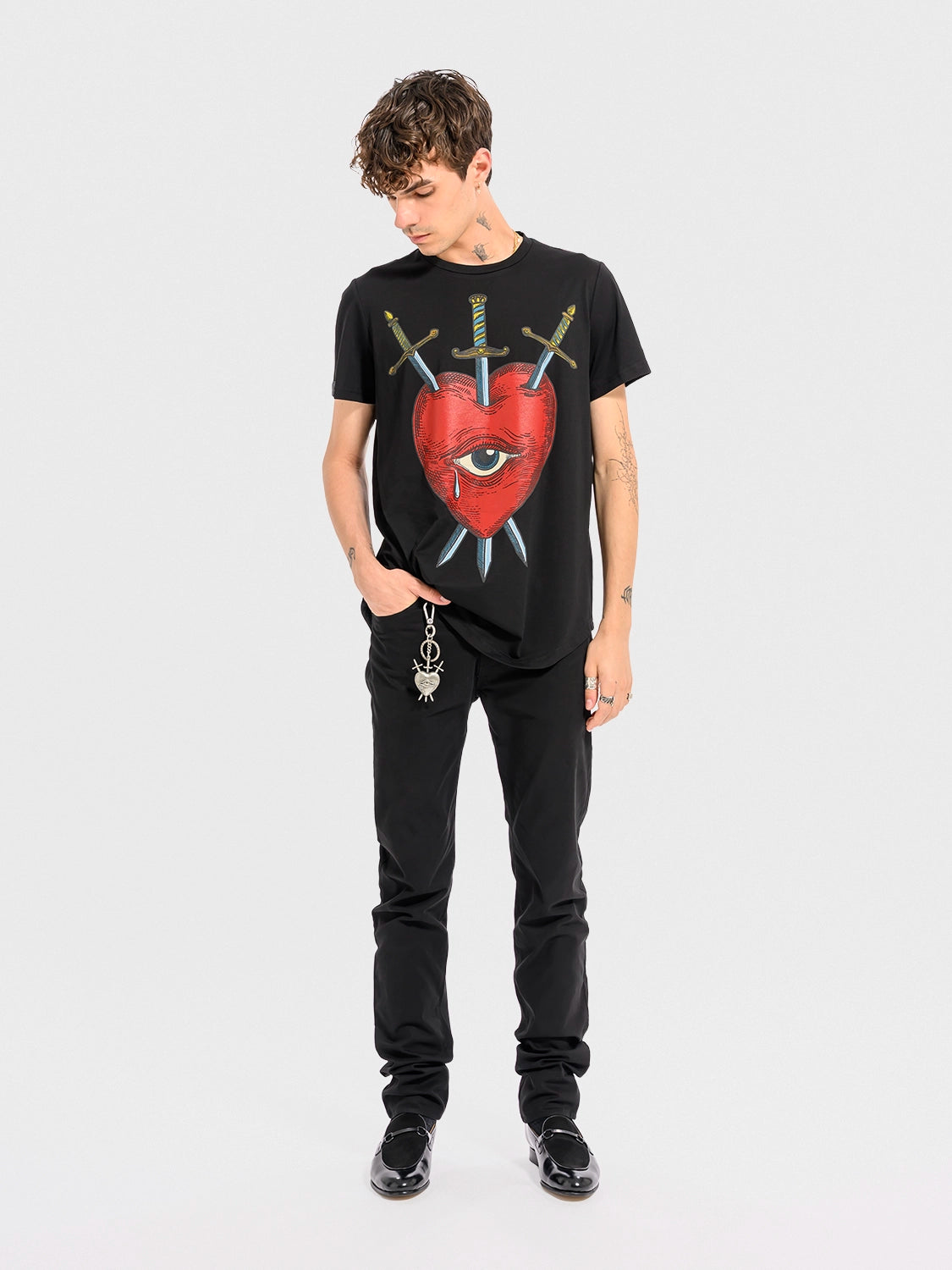 Person wearing a Black crew neck t-shirt with a graphic print of a red heart, an eye, and crossed swords.