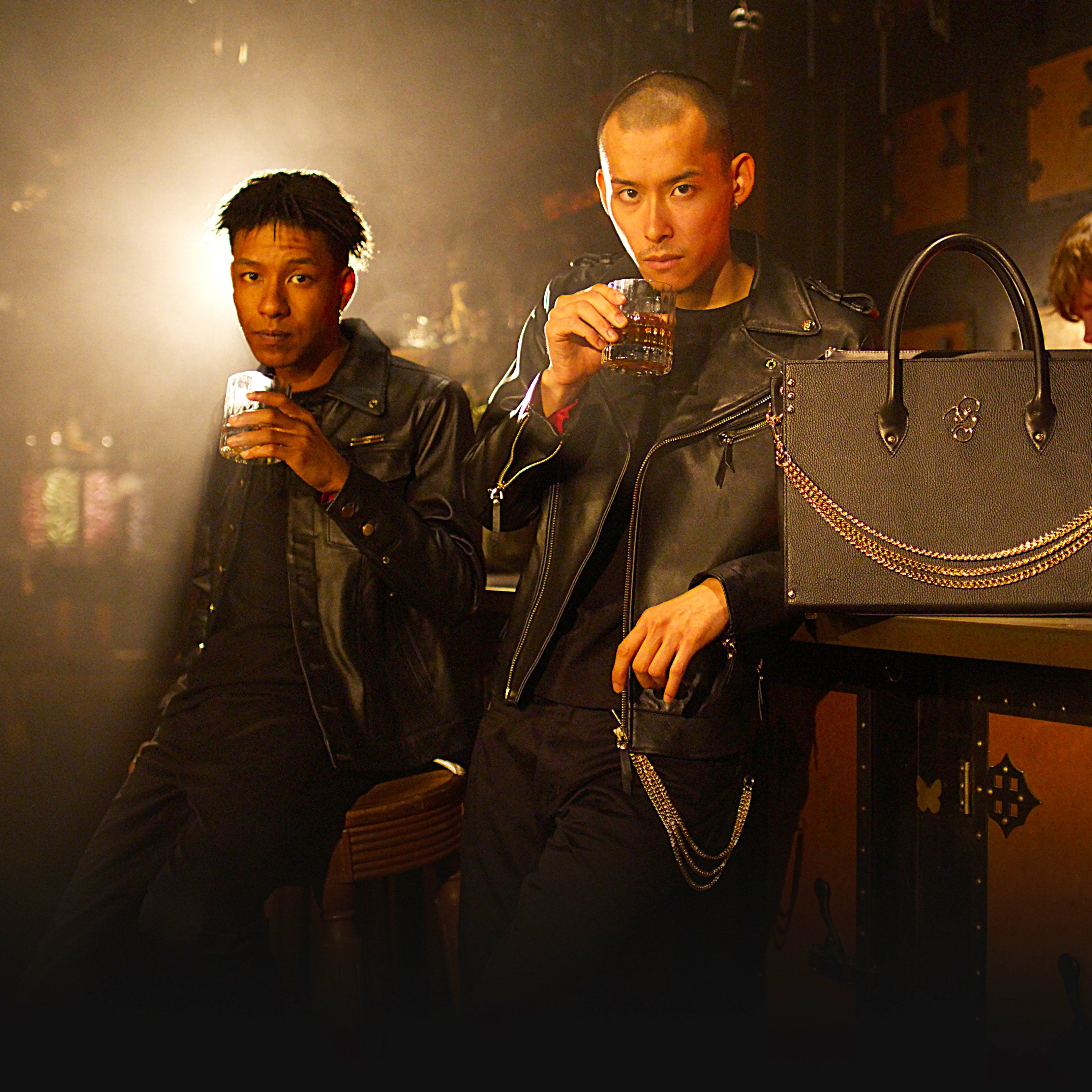 Two men dressed in black leather jackets holding glasses of whiskey in a bar setting, with a black bag placed on the counter beside them.