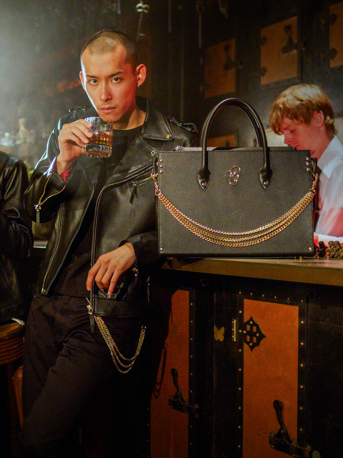 Man dressed in black with a black leather jacket, holding a glass of whiskey in a bar setting, with a black bag placed on the counter beside him.