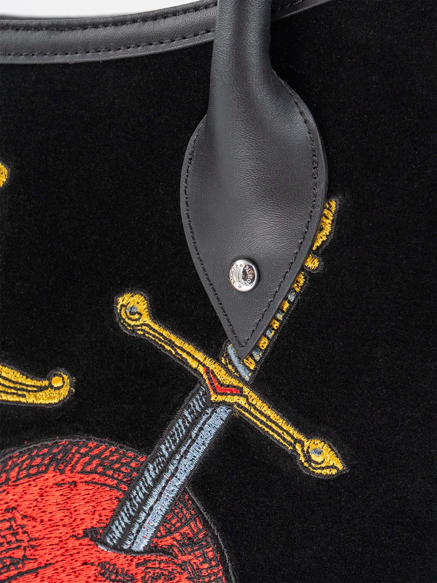 Close up of a black velvet and leather tote bag with a red heart, an eye, and crossed swords embroidered on the front, with a black handle.
