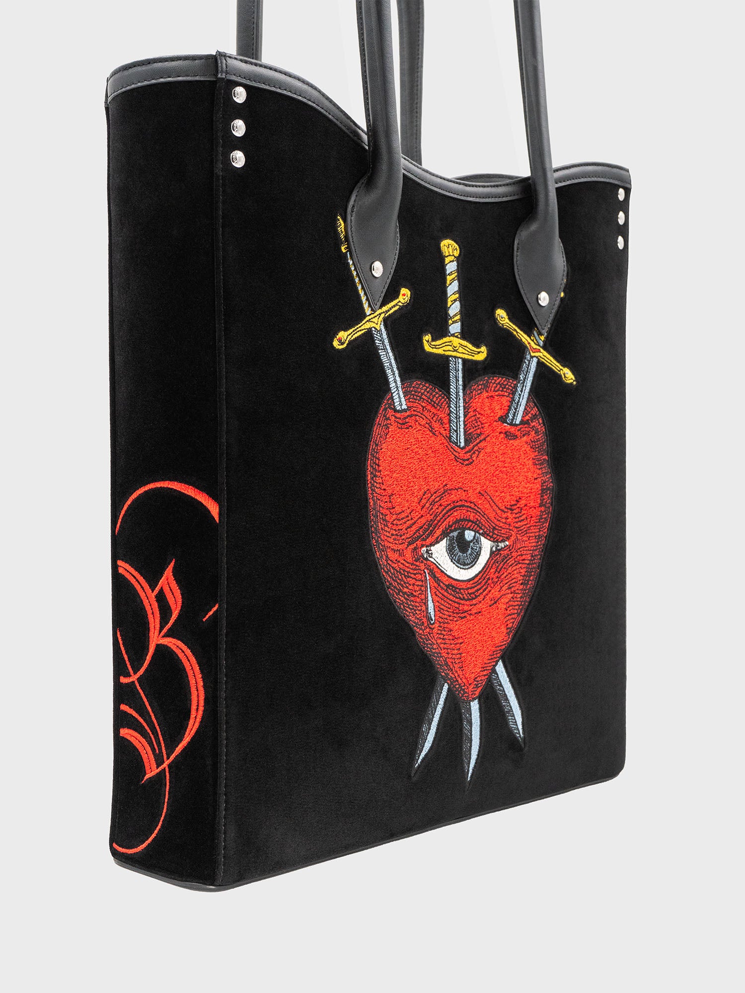 Side view of a black velvet and leather tote bag with a red heart, an eye, and crossed swords embroidered on the front, with a black handle.