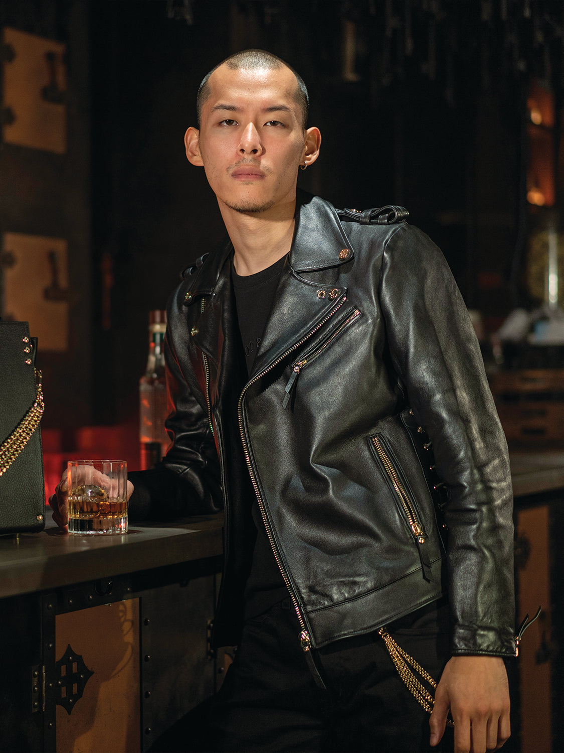 Man dressed in black with a black leather jacket, holding a glass of whiskey in a bar setting