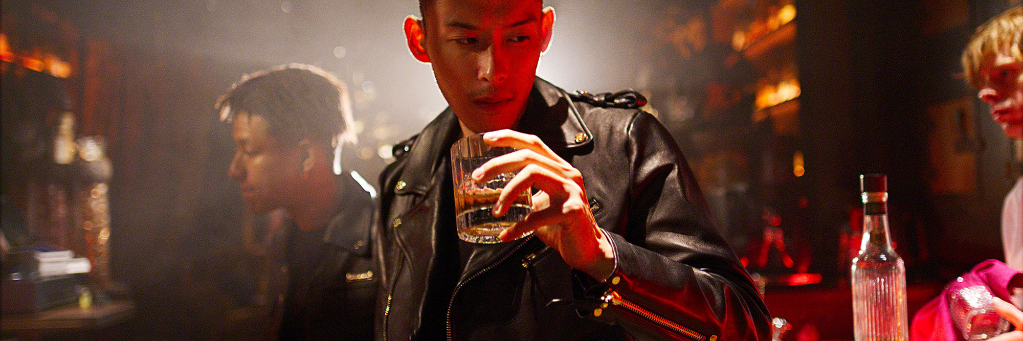 Person in a leather jacket holding a drink in a bar setting.