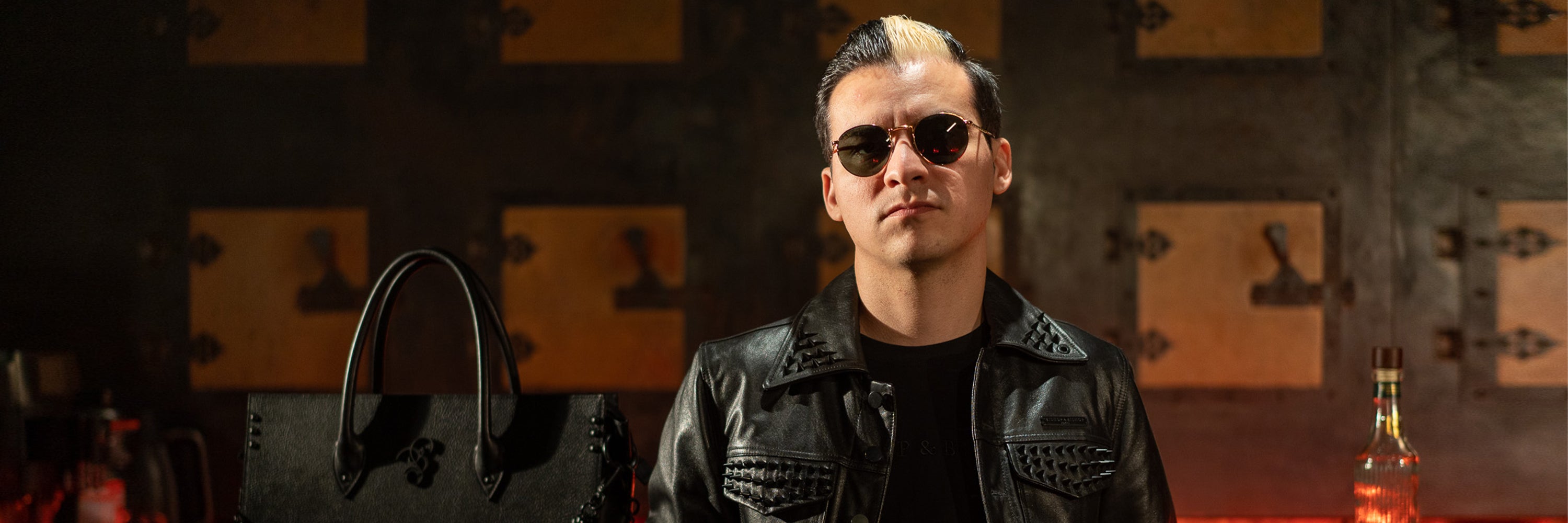 Close-up of Erik wearing sunglasses and a black leather jacket in a bar setting, with a black bag next to him.
