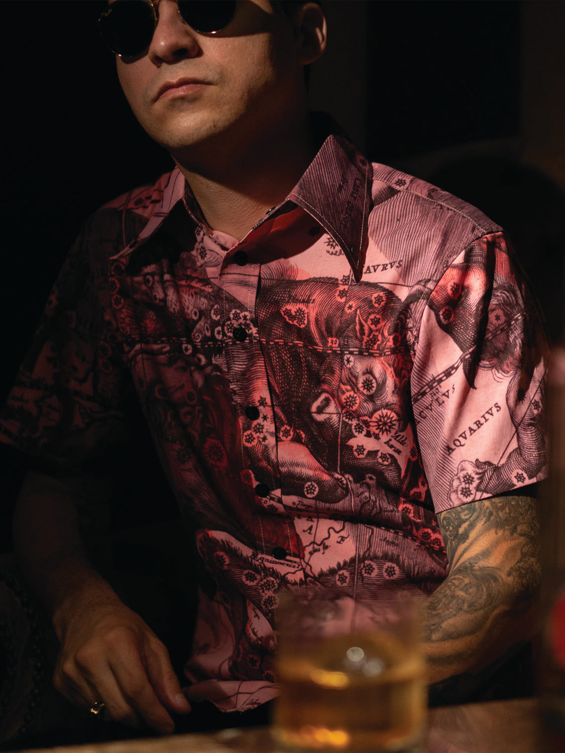 Man wearing a pink graphic shirt and sunglasses, sitting with a glass of whiskey in front of him.