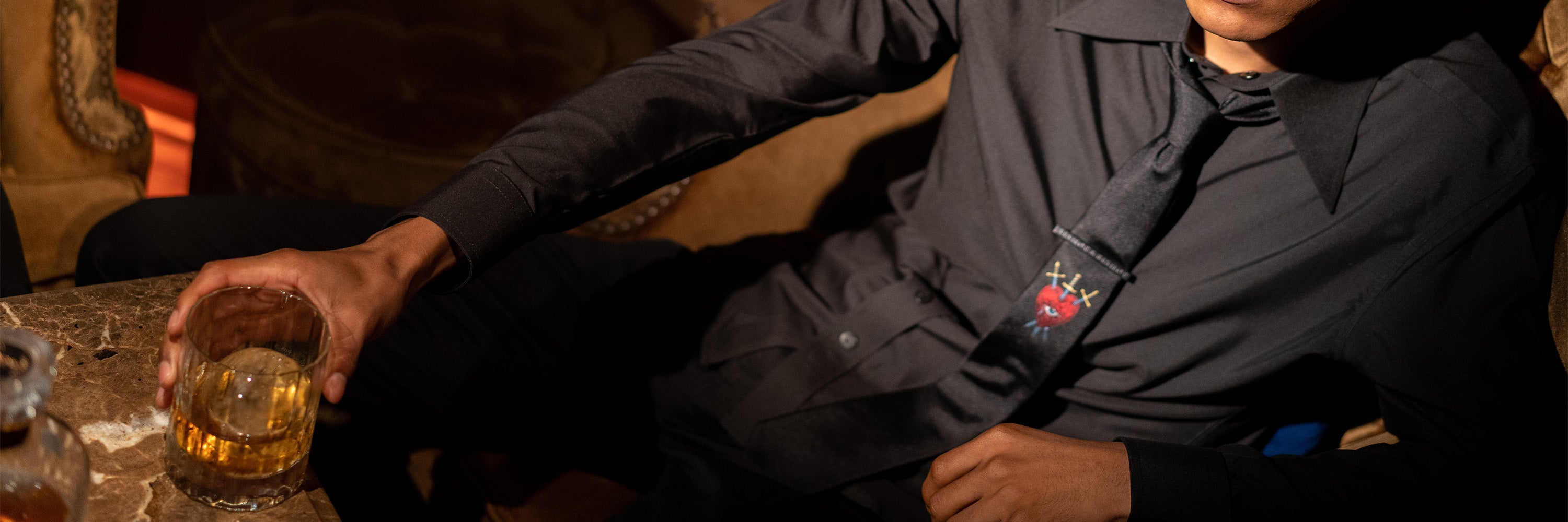 Man wearing a black shirt, black trousers, and a black tie with a red heart and crossed swords graphic, sitting in an armchair and holding a glass of whiskey, with his head out of frame.