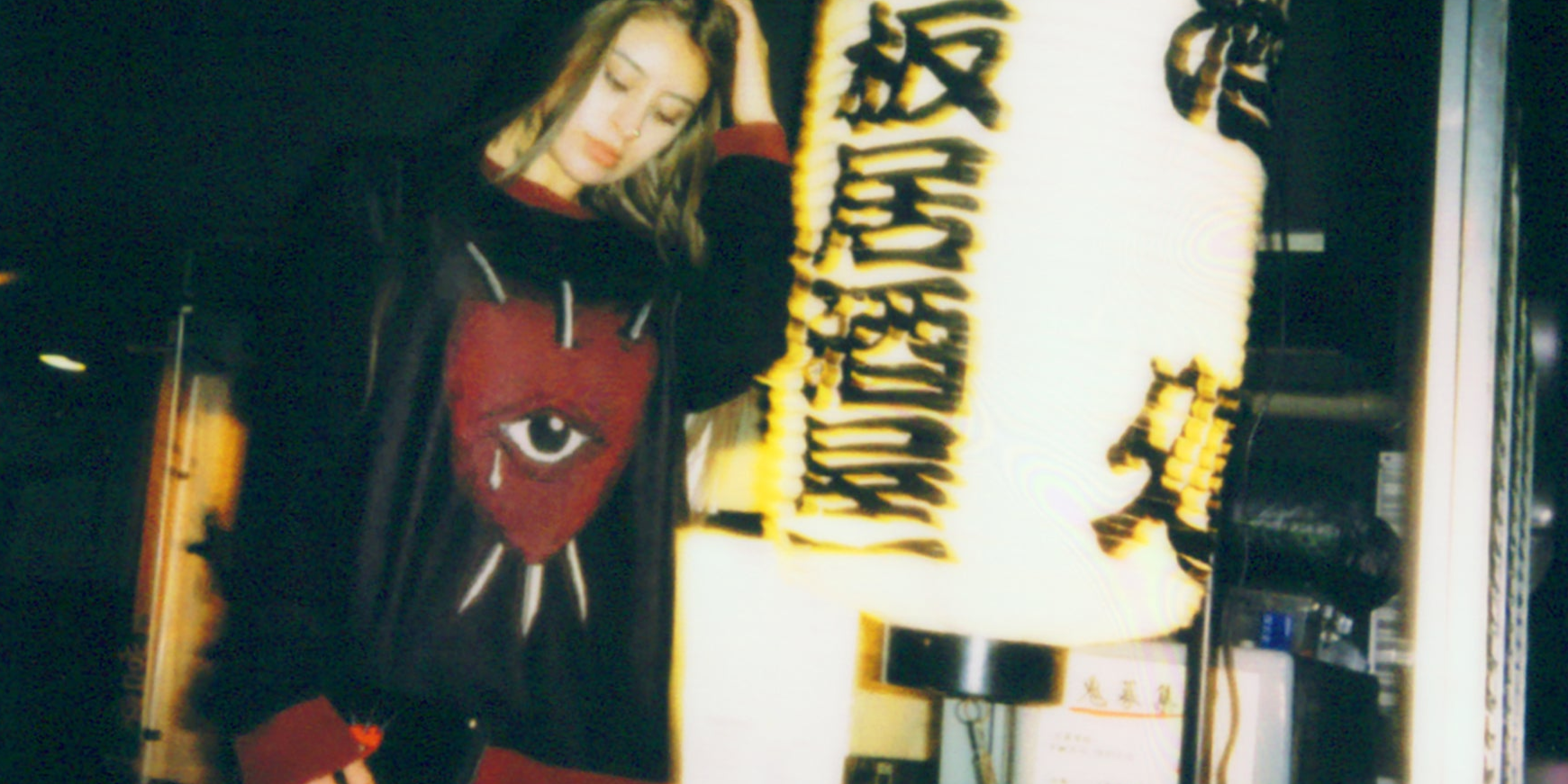 Woman wearing a black sweater featuring a red heart with an eye pierced by three swords graphic, photographed outdoors.