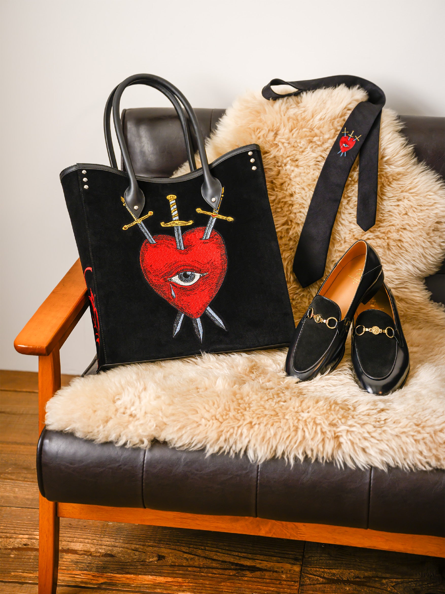 Black bag featuring a red heart with an eye pierced by three swords graphic, placed on a fur-covered armchair with a matching tie draped over the backrest and black shoes with gold detailing beside it.