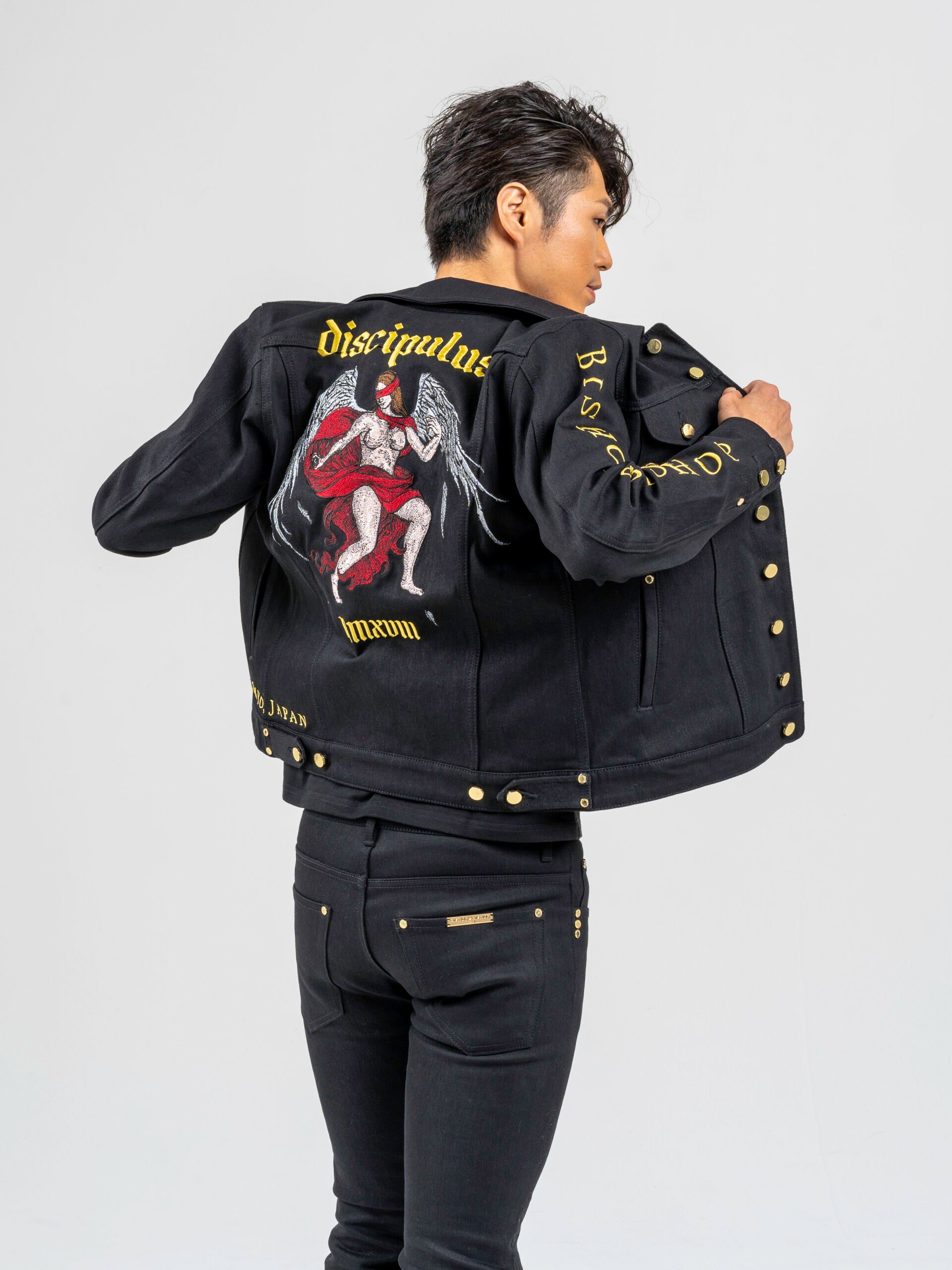 Rear view of a man wearing the black trucker jacket, showcasing gold Japanese embroidery of an angel with wings and a red blindfold across the eyes.