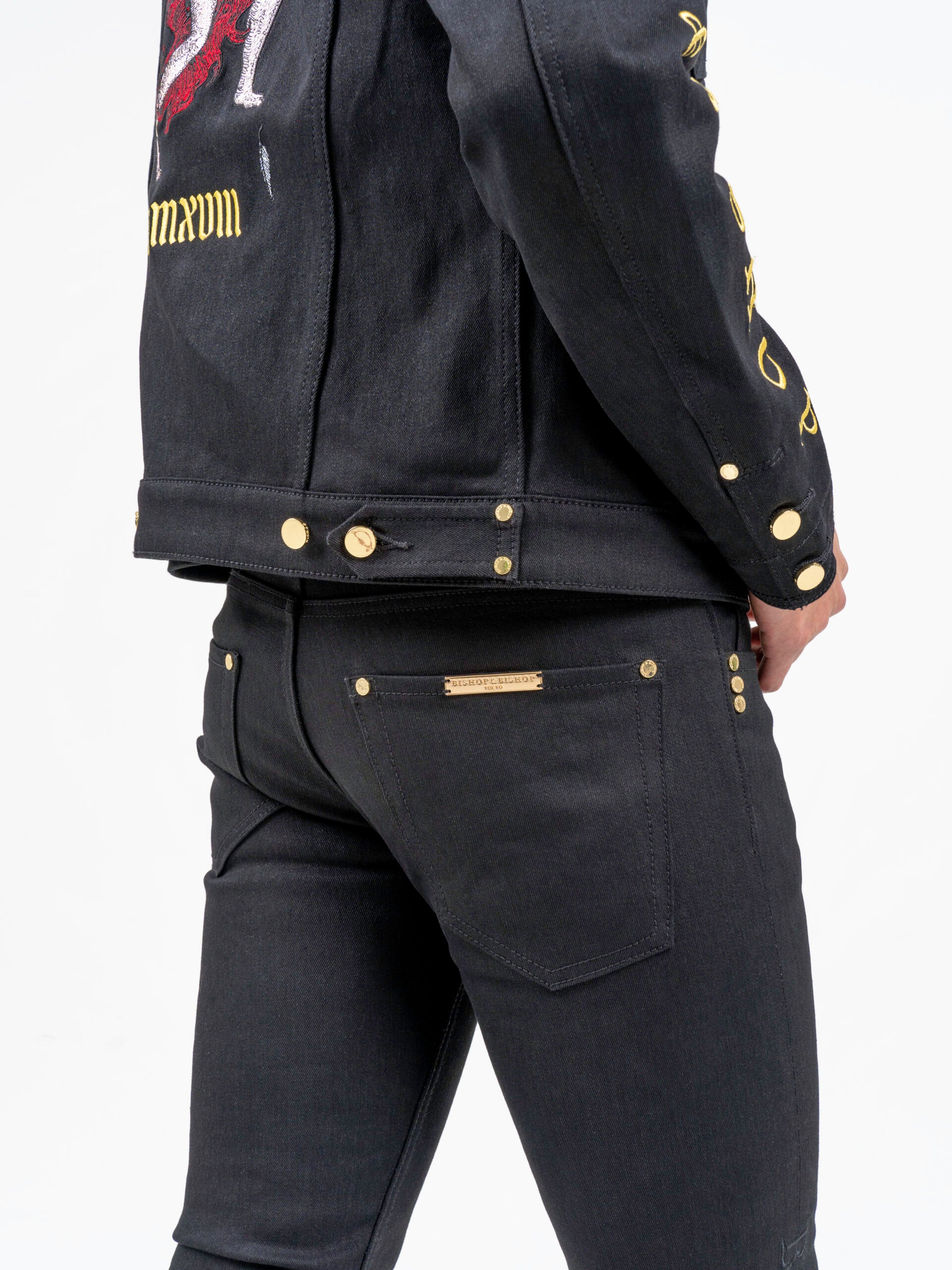 Person wearing a black jacket and black trousers, shown from the back, with a faint angel graphic visible on the jacket, and gold buttons on both the jacket and trousers.
