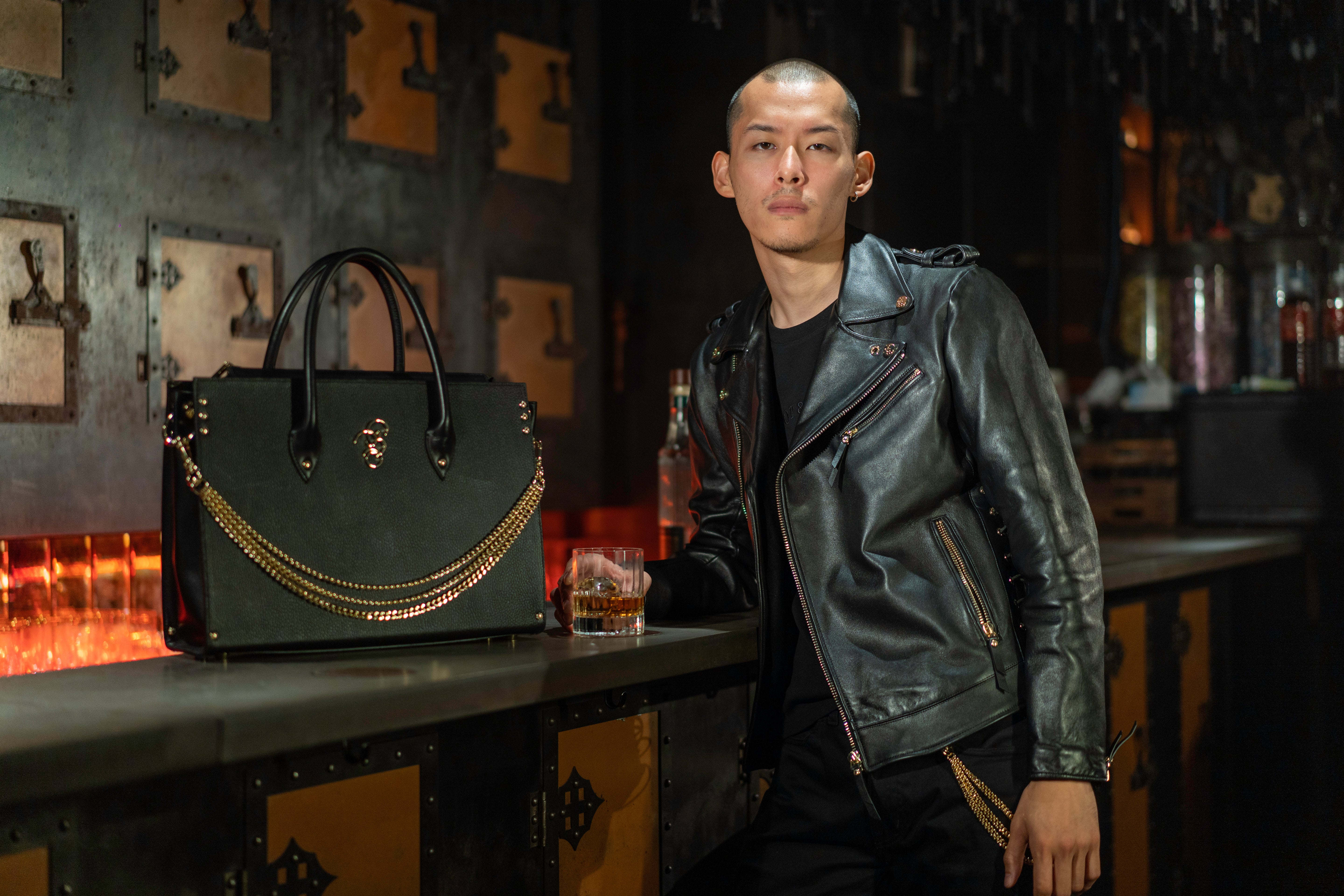 Man wearing a black leather jacket and black trousers, standing at a counter with a glass of whiskey and a black bag.