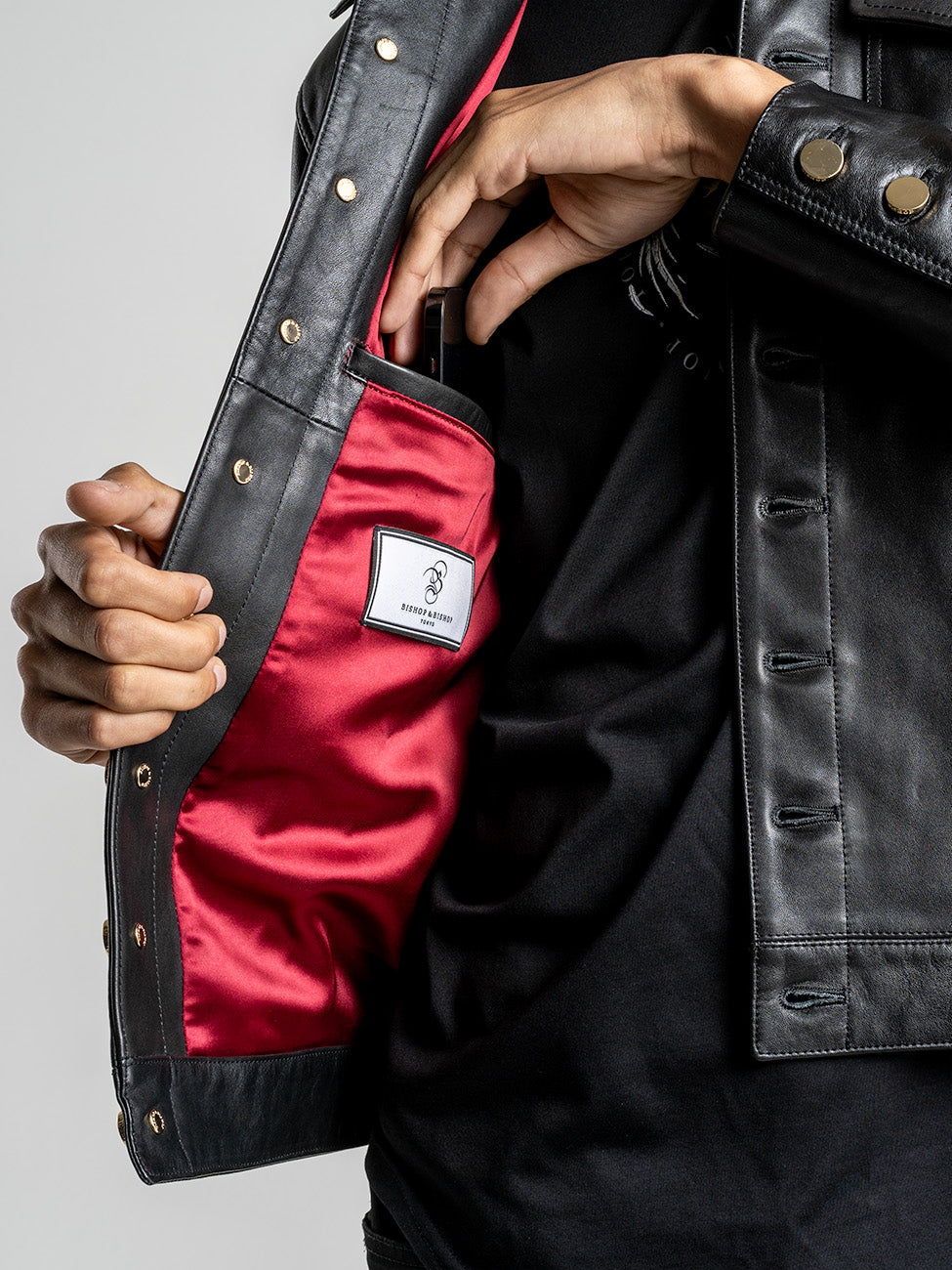 Close-up of an open black leather jacket showing an interior pocket, red lining, and a label reading “Bishop & Bishop Tokyo.”