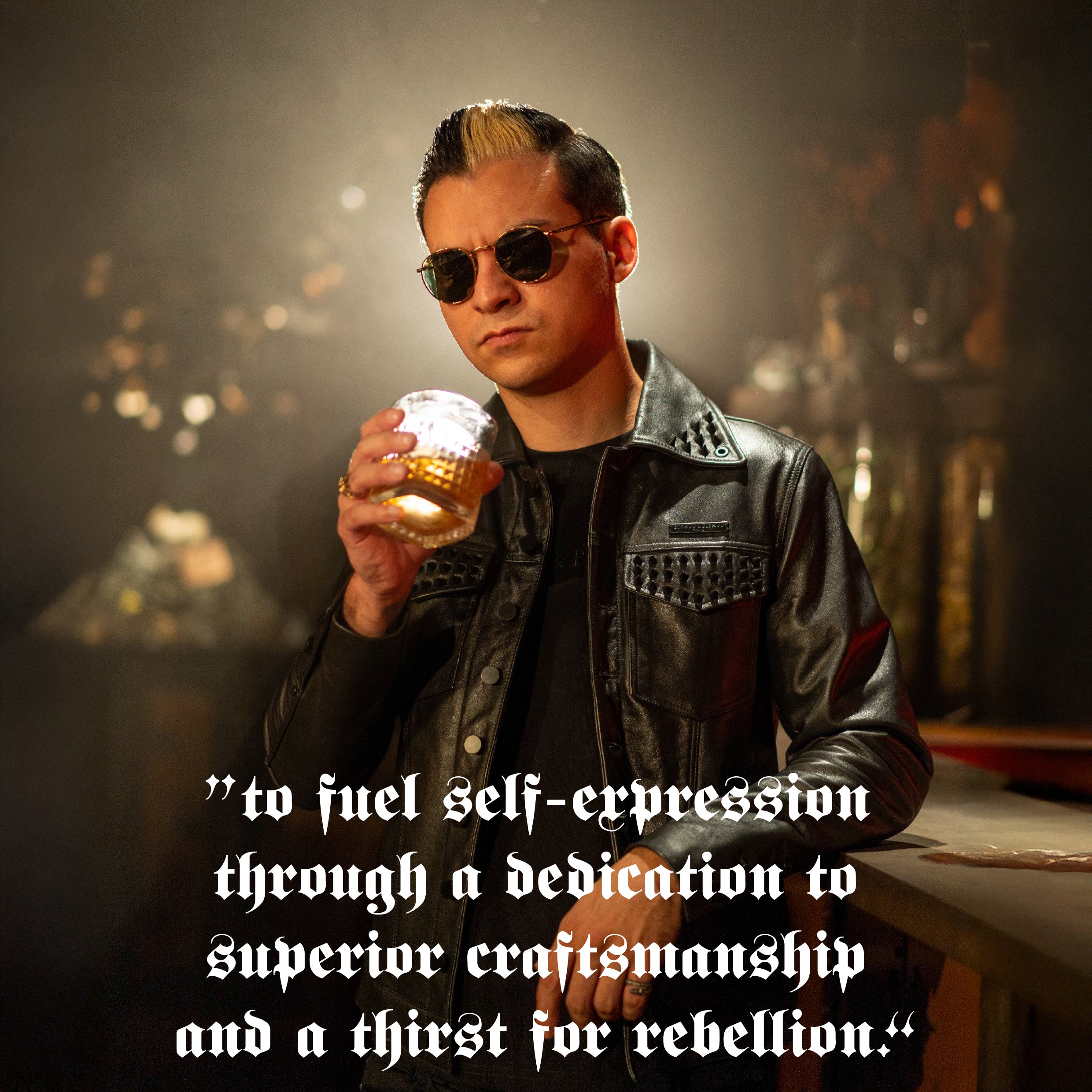 Erik wearing a leather jacket and sunglasses, holding a glass of whiskey, with overlaid text reading “to fuel self expression through a dedication to superior craftsmanship and a thirst for rebellion.”