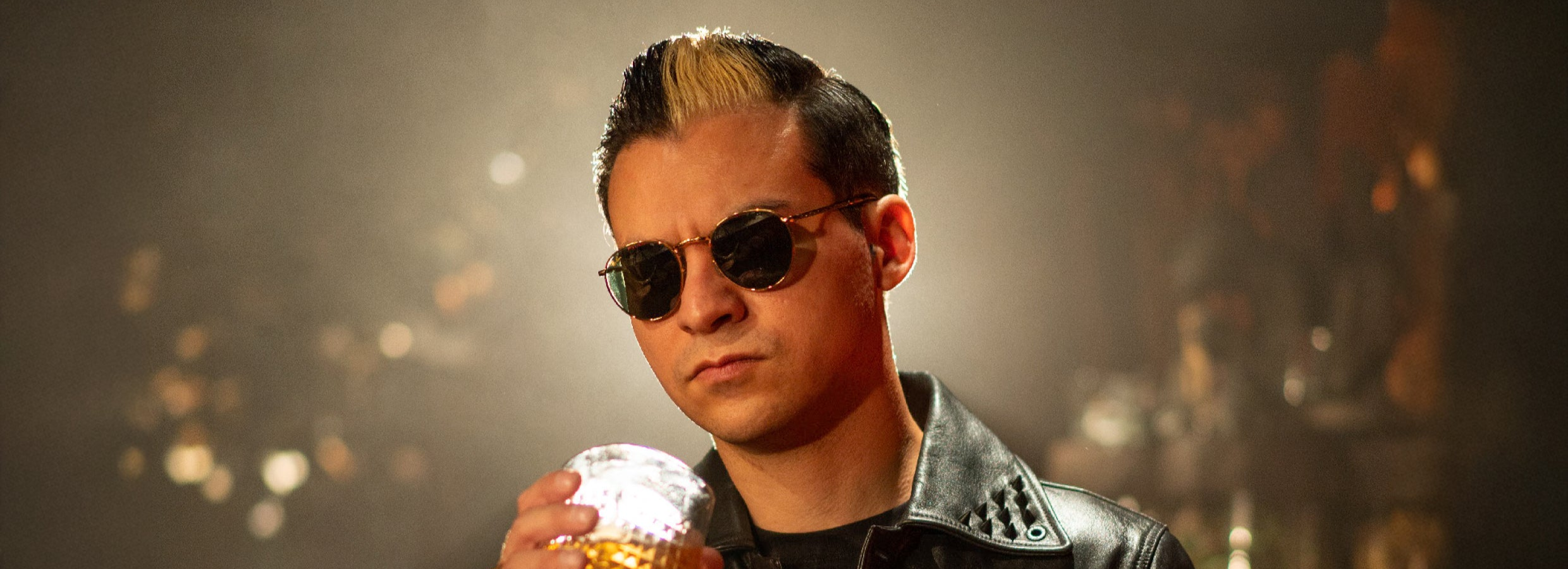 Close-up of Erik wearing a leather jacket and sunglasses, holding a glass of whiskey.