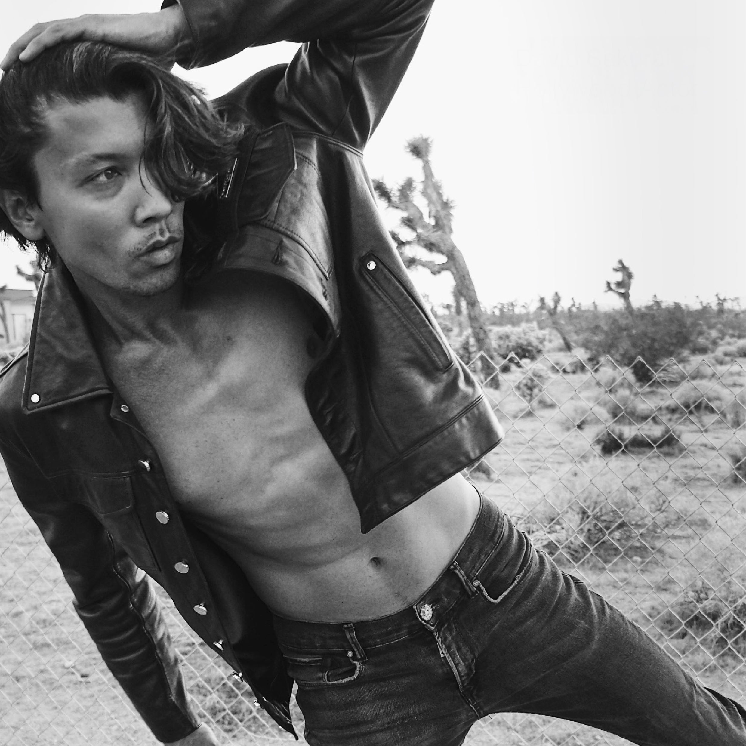 Grey-tone image of David Sakurai wearing the custom black leather trucker jacket with no shirt underneath, highlighting its clean design, premium leather texture, and bold, minimalist silhouette.