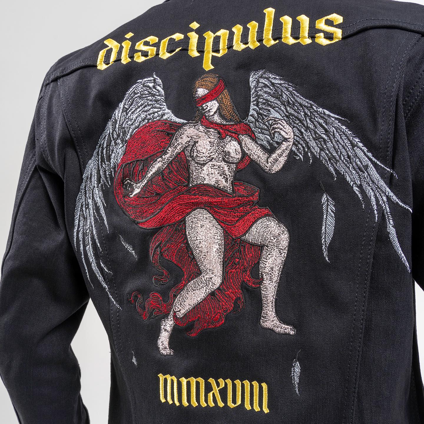 Rear view of a jacket featuring embroidery of an angel with wings, a red blindfold, and a flowing red cloak, with “DISCIPULUS” embroidered above and “MMXVIII” below.