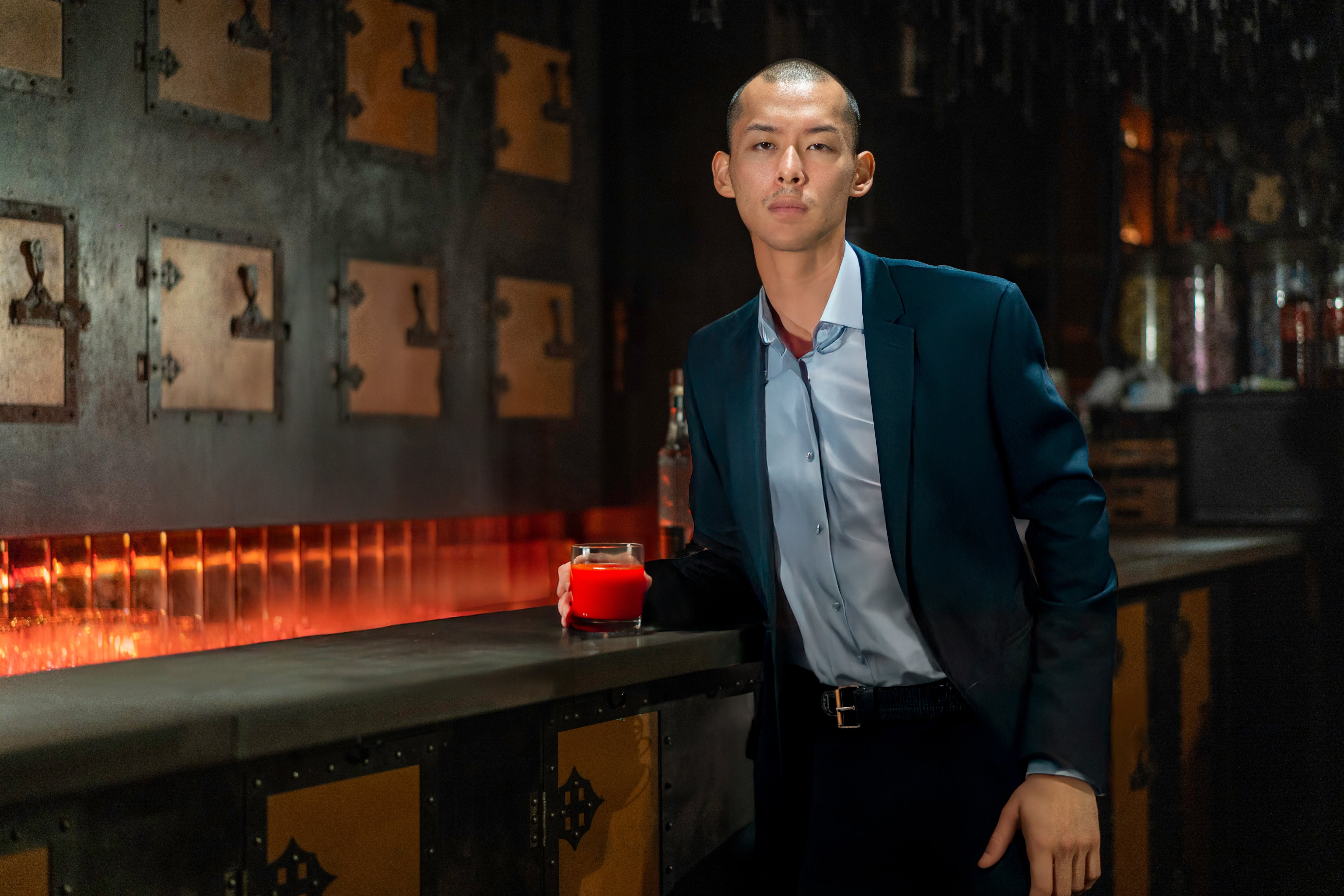 Man wearing a blue blazer, light blue shirt, and black trousers, standing at a counter with a glass of bright red drink.