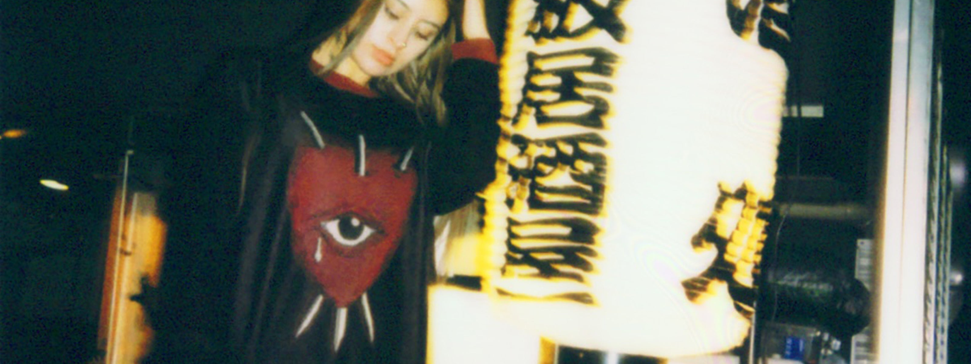 Woman wearing a black sweater featuring a red heart with an eye pierced by three swords, photographed in an indoor setting.