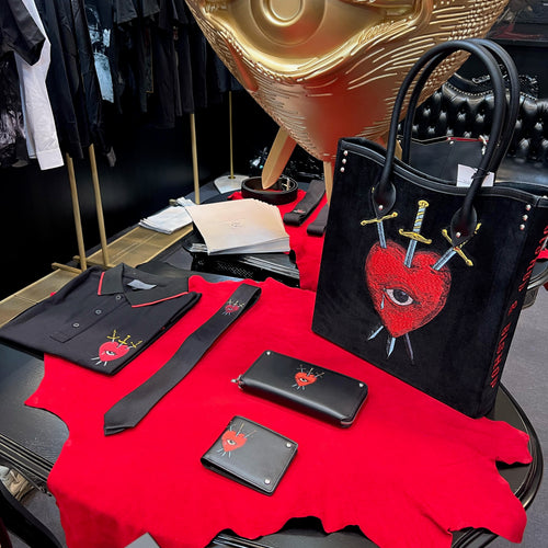 Table covered with red fabric displaying a black handbag, wallets, a T-shirt, and a tie featuring a red heart with an eye pierced by three swords, with a gold eye sculpture in the background.