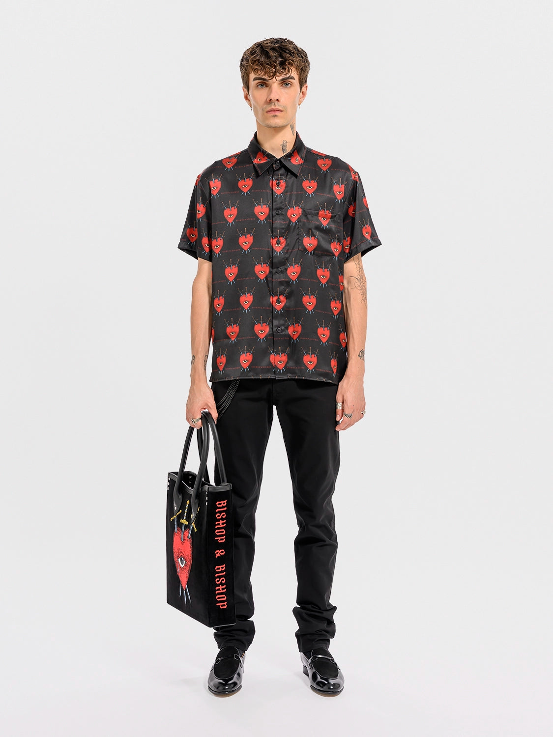 Person wearing a black shirt with  a red heart, an eye, and crossed swords patterns, holding a black bag with  a red heart, an eye, and crossed swords designs on a white background.