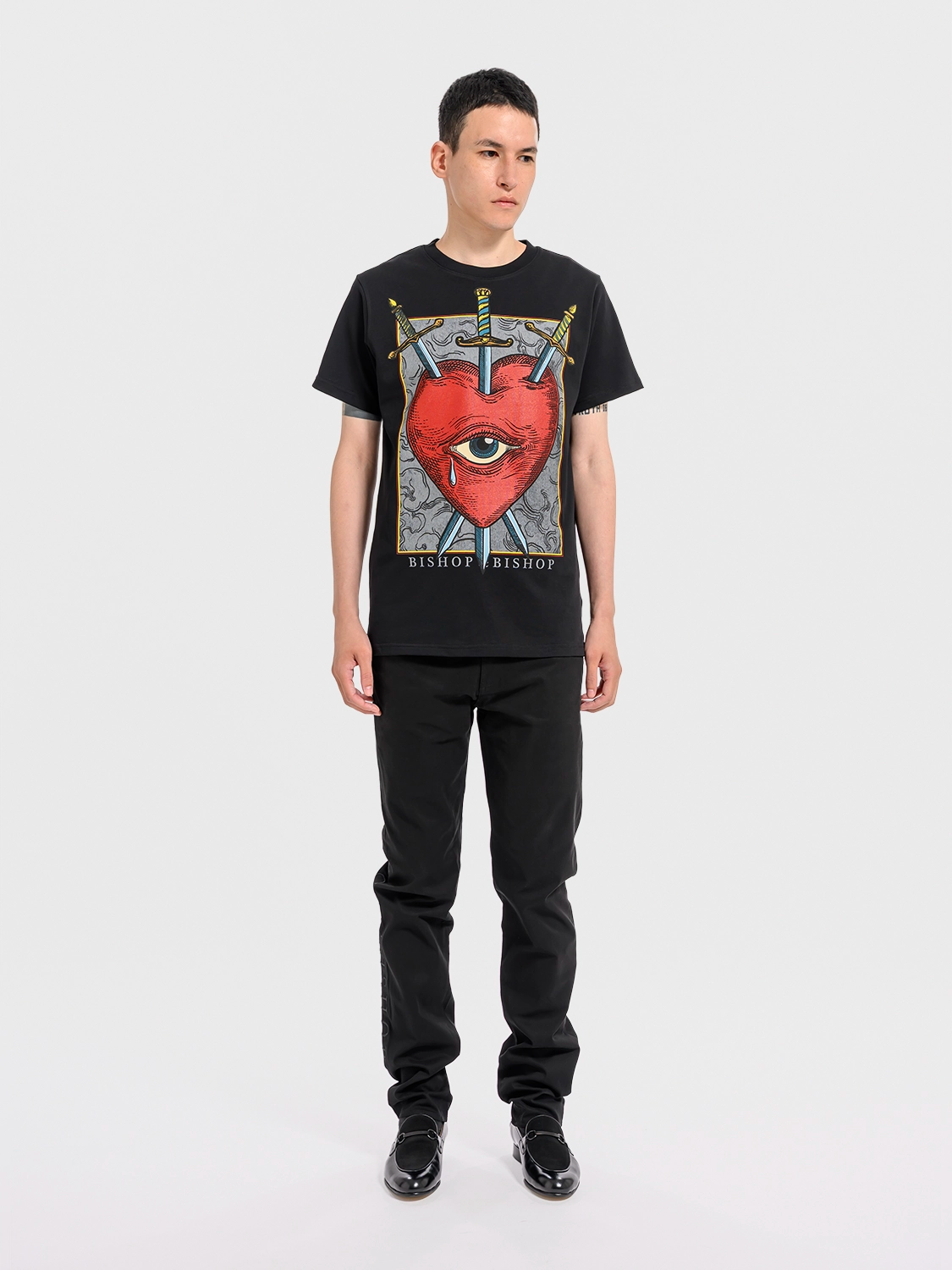 Person wearing a black crew neck t-shirt with a graphic print of a red heart, an eye, and crossed swords, and the text 'BISHOP BISHOP'.