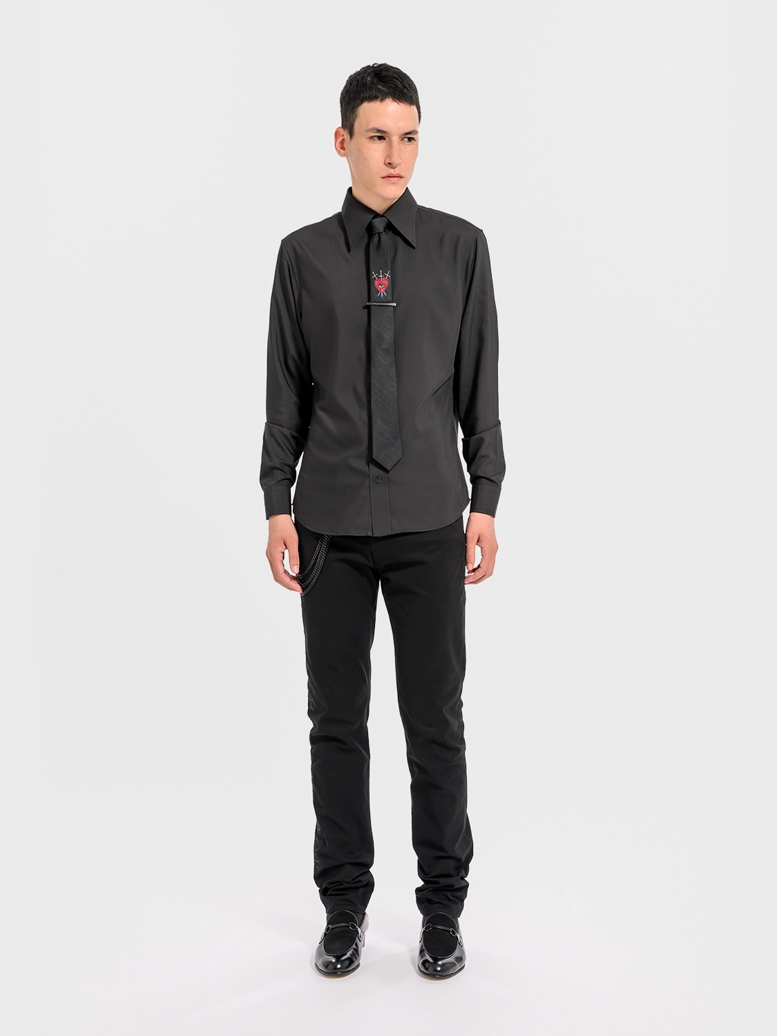 Person wearing a black dress shirt with a black tie with a red heart, an eye, and crossed swords and black pants on a white background